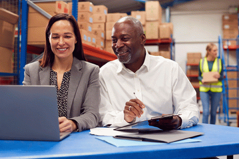 Two individuals are sitting at a blue table in a storage room with numerous brown packaging boxes stacked on shelves in the background. On the left is a Black male with short grey hair and matching facial hair. He wears a white long-sleeved dress shirt and a silver ring on his left hand. He holds a pen in his left hand and a clipboard in his right hand. He smiles as he looks at a grey laptop on the table. On the right is a White female with long, straight brown hair. She wears a grey blazer.